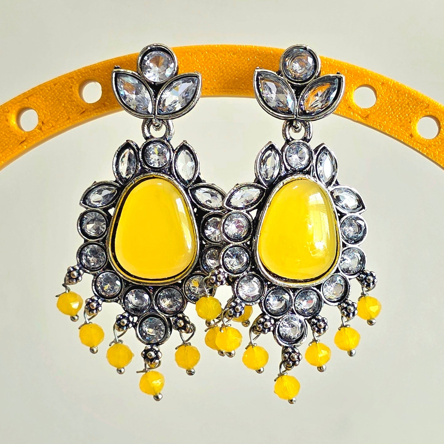 Yellow earrings with silver and crystal details on a yellow stand against a light gray background