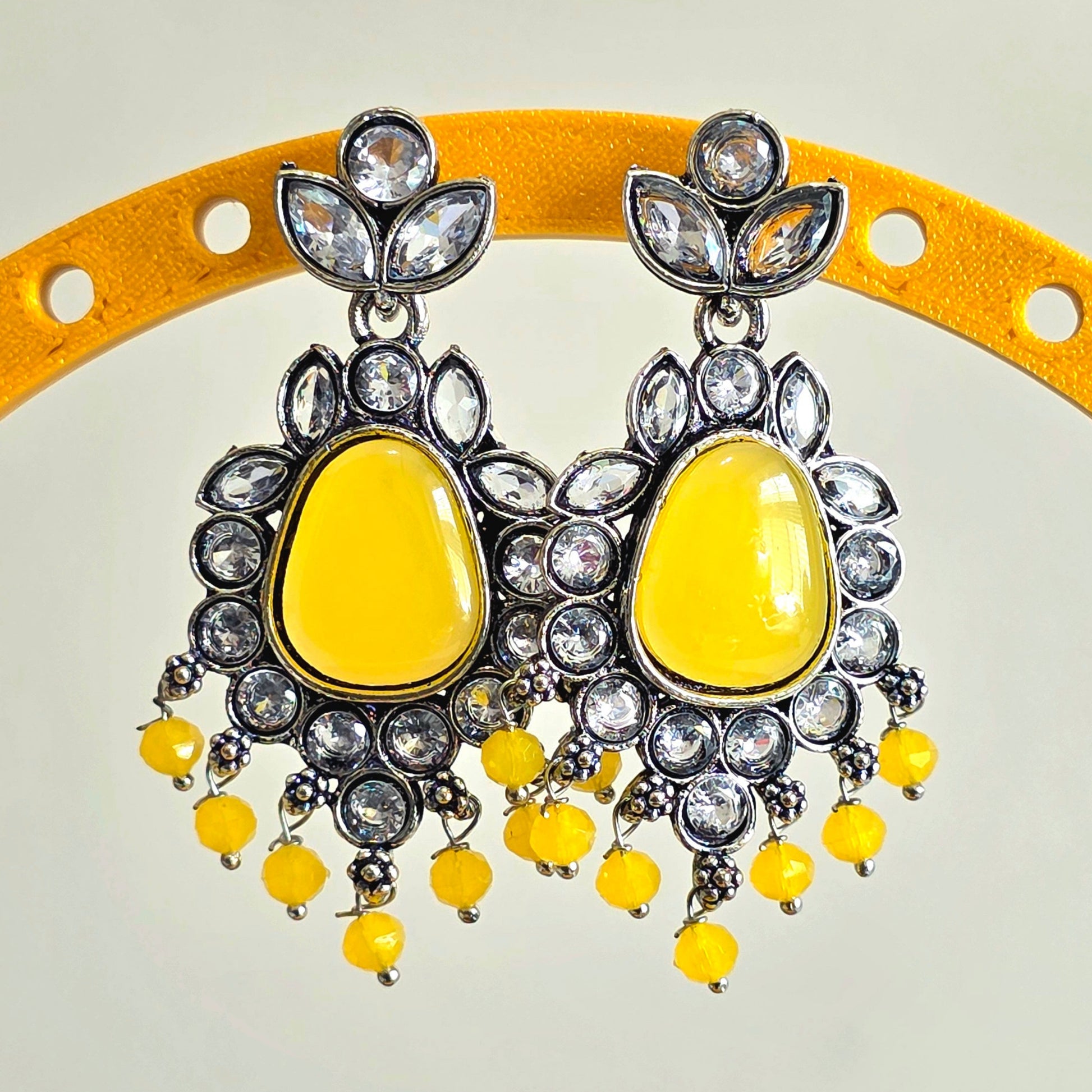 Yellow earrings with silver and crystal details on a yellow stand against a light gray background