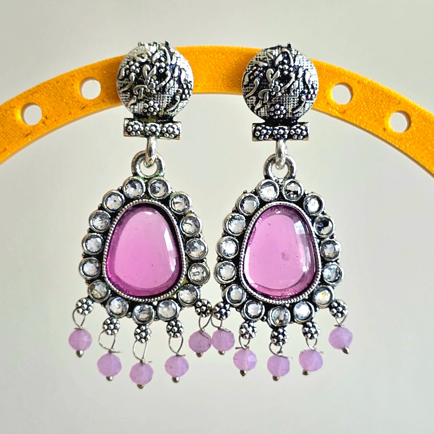Pink earrings with silver and crystal details on a yellow stand against a white background